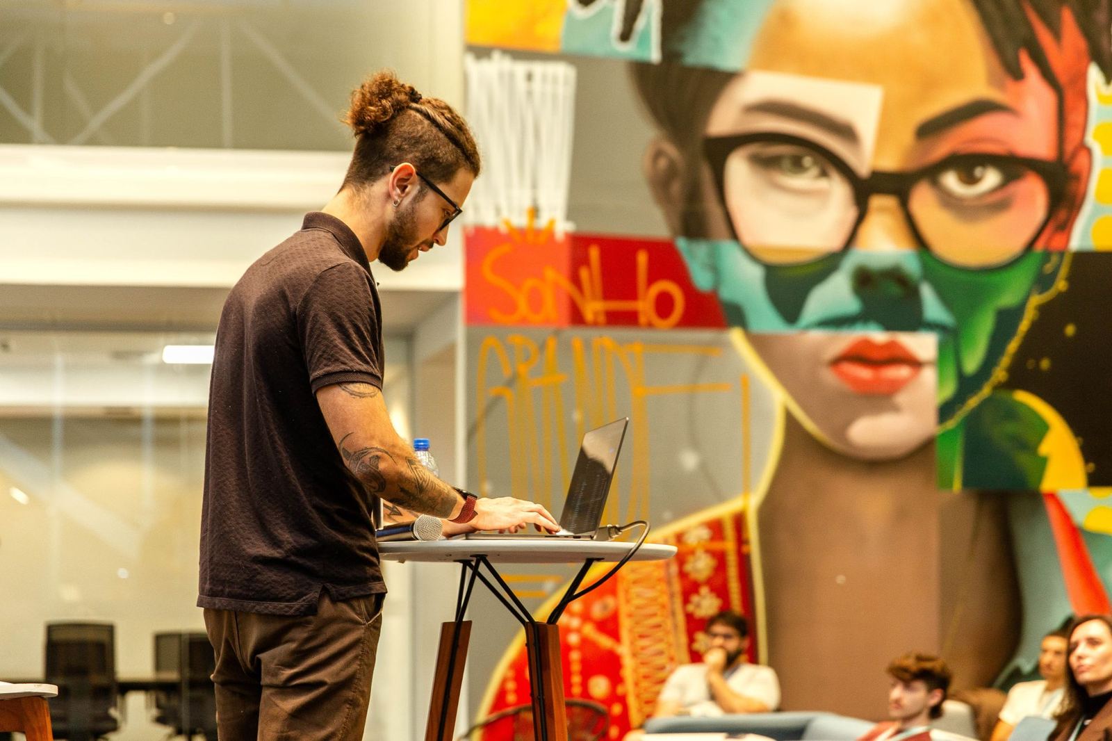 Matheus Kemer de perfil, em pé diante de um notebook, com um grande mural colorido com os dizeres “SONHO GRANDE” ao fundo.