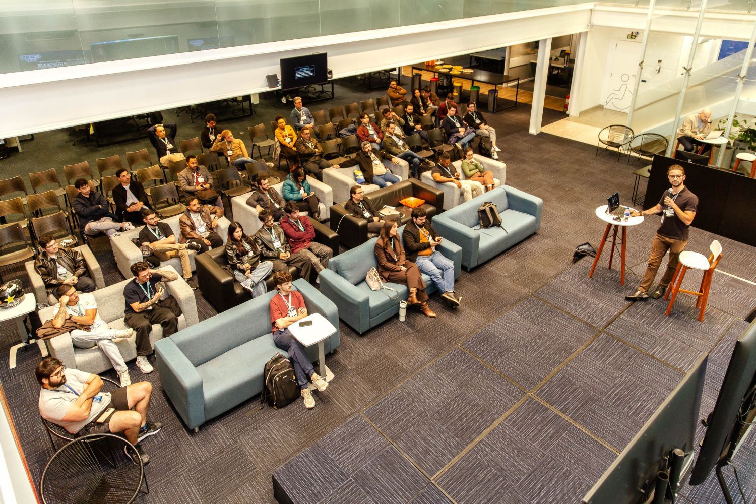 Vista do alto do auditório do AI Tinkerers Curitiba: cerca de 40 pessoas sentadas em sofás e poltronas, com Matheus Kemer apresentando à direita.